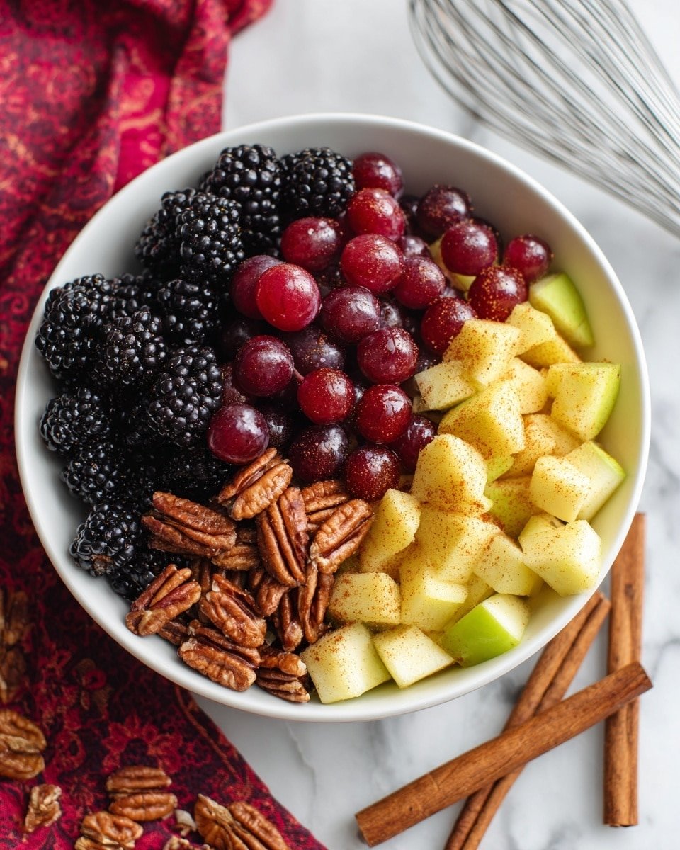 A white bowl filled with a fresh fruit mix showing three main layers: the bottom layer has blackberries, the middle layer has red grapes, and the top layer consists of light yellow apple pieces sprinkled with cinnamon and mixed with small brown pecans. The bowl sits on a white marbled surface with two cinnamon sticks and a metal whisk nearby, with part of a red patterned cloth in the background. The fruits look juicy and the cinnamon gives a warm color touch to the apples and pecans photo taken with an iphone --ar 4:5 --v 7 — Fall Fruit Salad with Citrus Maple Cinnamon Dressing, autumn fruit salad, fall healthy recipes, fall brunch ideas, seasonal fruit salad